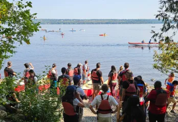 A group of people with red life jackets on. They are standing by red canoes on the Ottawa River shore and watching other people in canoes on the water.