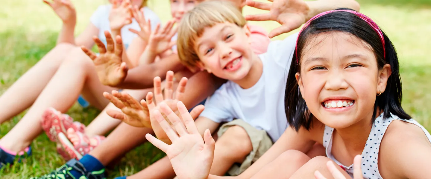 A group of children sit in a row smiling and waving.