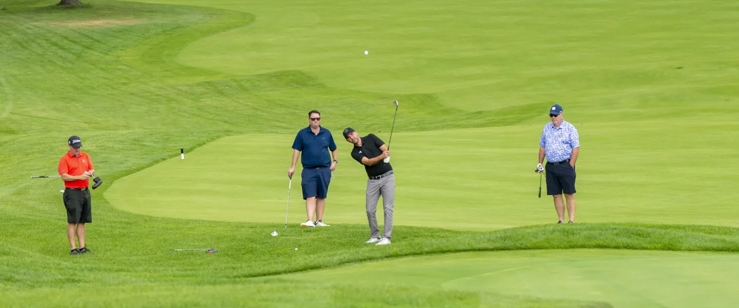 A group of four people holding clubs on a golf course. One is swinging their club.