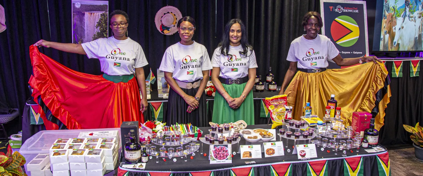 Guyana table covered in food and decorated with flags. Four women are standing behind the table, their skirts the colours of the Guyana flag.