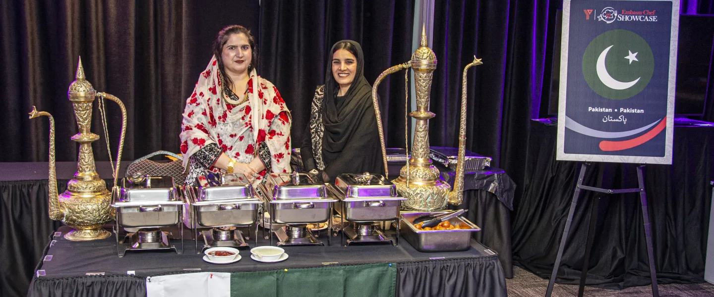 Two women in traditional attire at a Pakistan table covered with food and a flag.