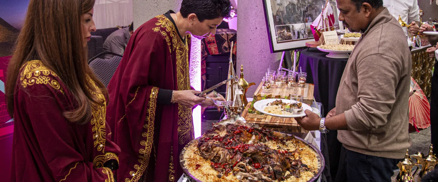 Two women in traditional attire serving Qatari food at a table.