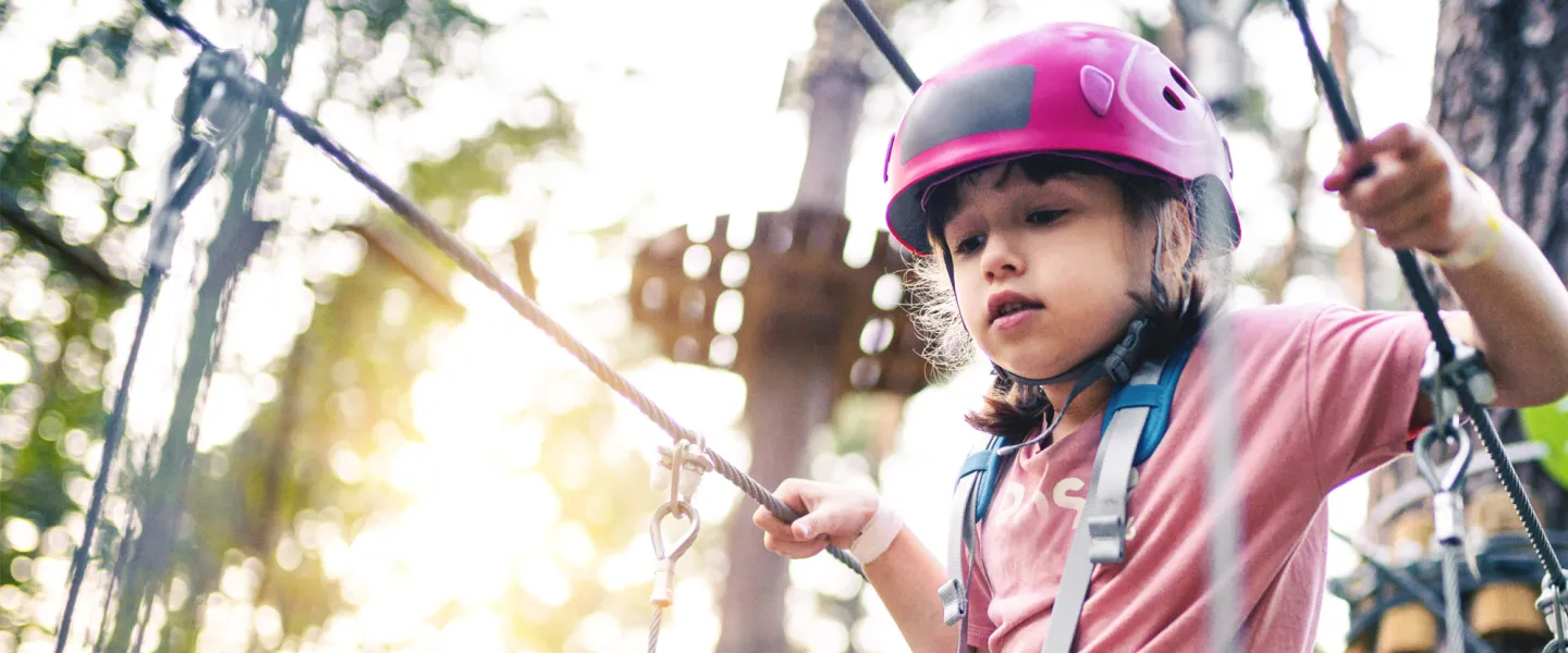 A girl wearing a helmet and safety harness navigates a high ropes course.