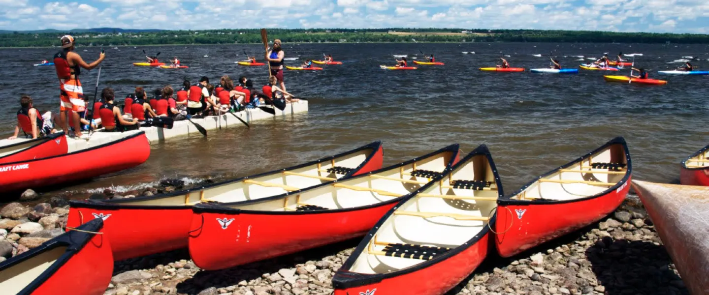 Red canoes sit on the shore in the foreground - in the background campers paddle in canoes.