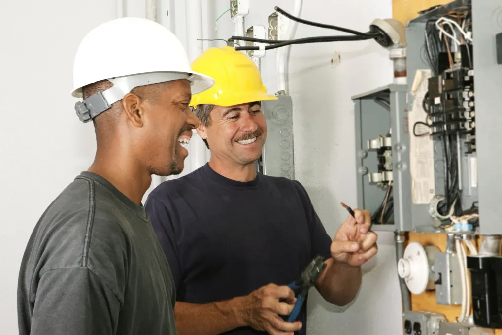 Two smiling electricians wearing hard hats working together in front of an electrical panel.