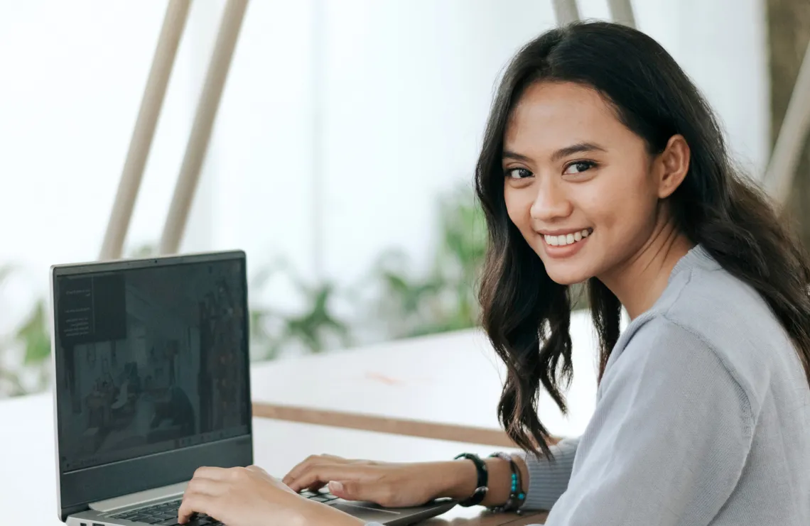 A young Asian woman smiles while working on her laptop.