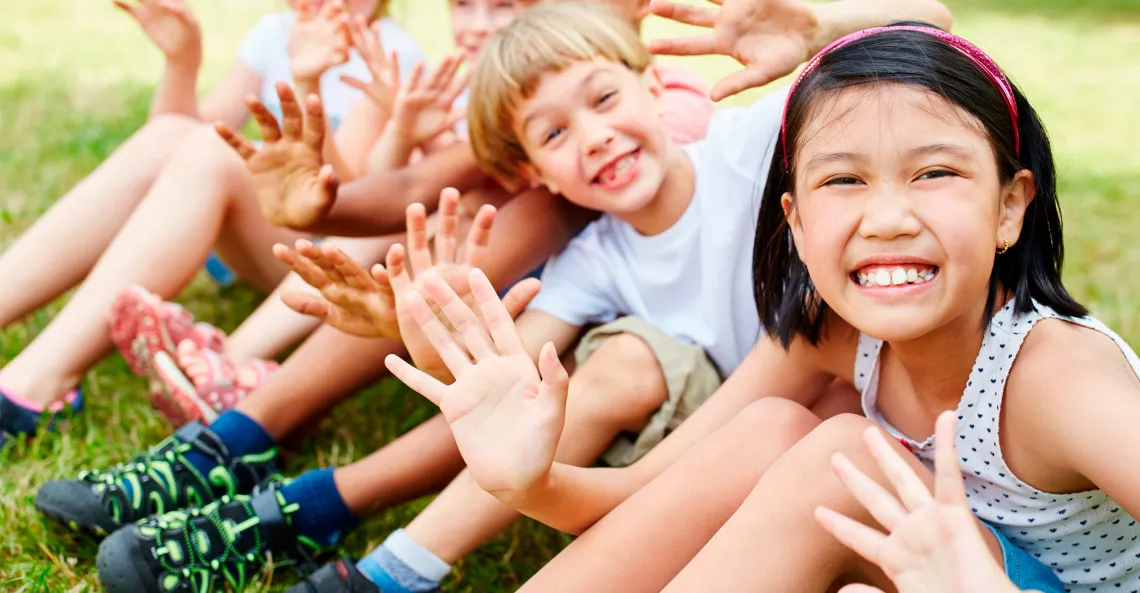 A group of children sit in a row smiling and waving.