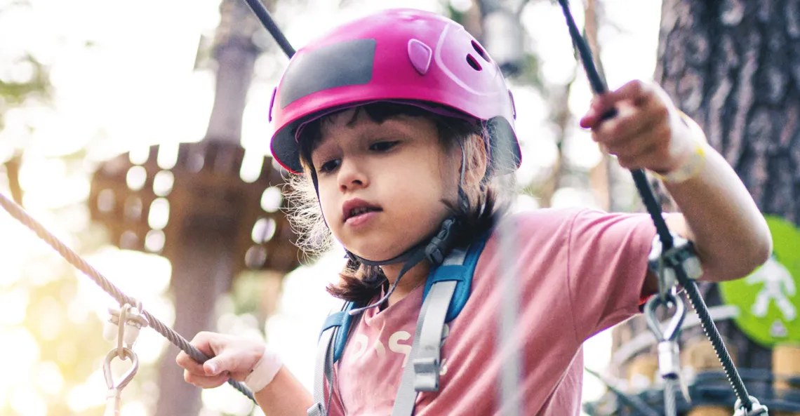 A girl wearing a helmet and safety harness navigates a high ropes course.